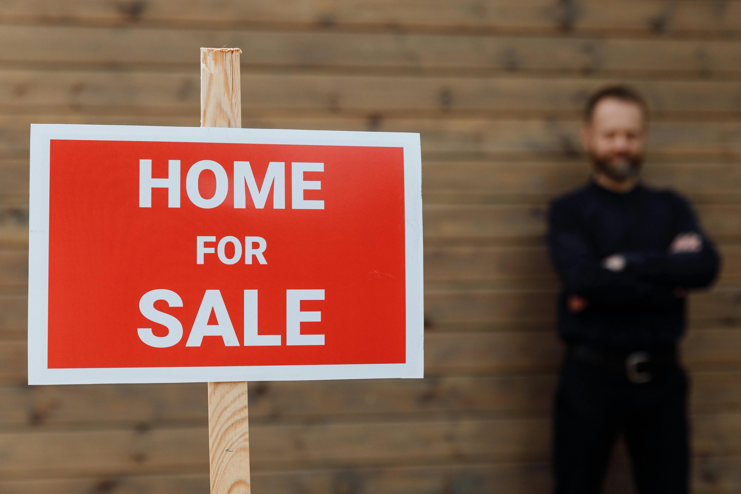 A confident real estate agent stands by a home for sale sign against a wooden wall.